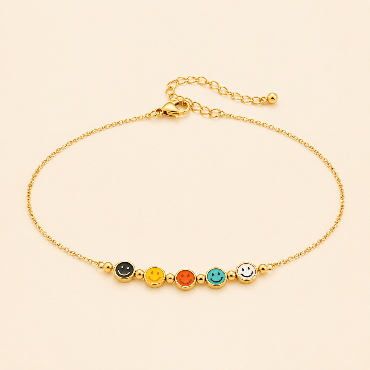 Gold anklet with five colourful enamel smiley face charms on a warm ivory background
